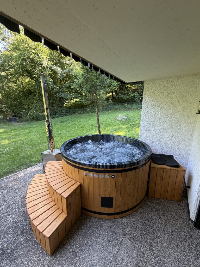 Wooden hot tub on a stone patio under a covered patio, with curved steps around the tub and greenery in the yard behind it.